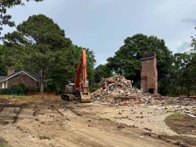 Excavator on a residential demolition site with debris pile and remaining brick chimney