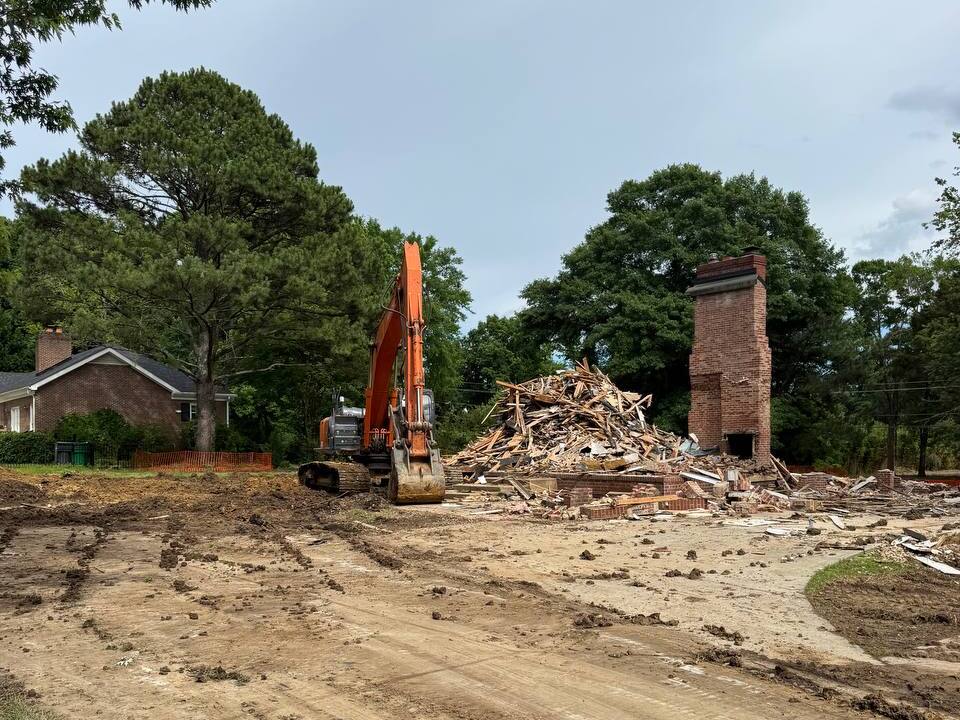 Excavator on a residential demolition site with debris pile and remaining brick chimney